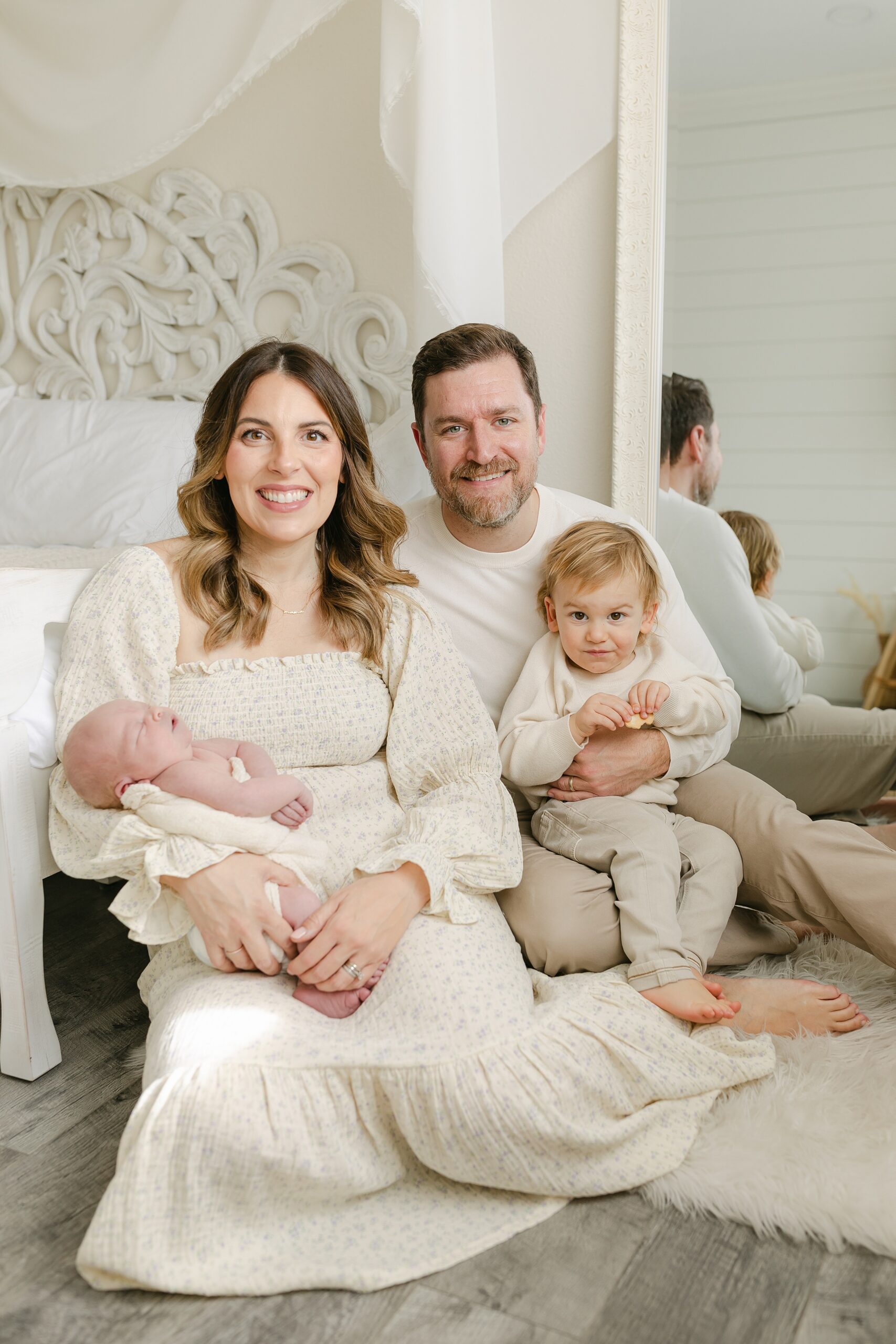 A mother and father sit on the floor of a studio against a bed with their newborn baby and toddler son