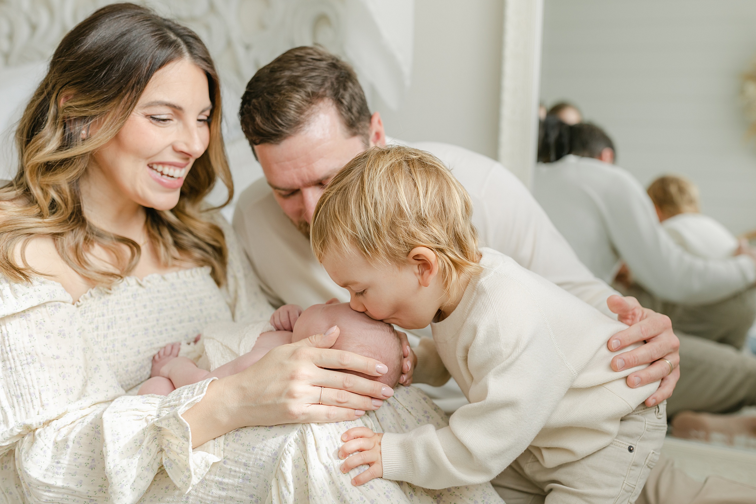 A toddler boy kisses the head of his newborn baby sibling in mom's lap while on a bed