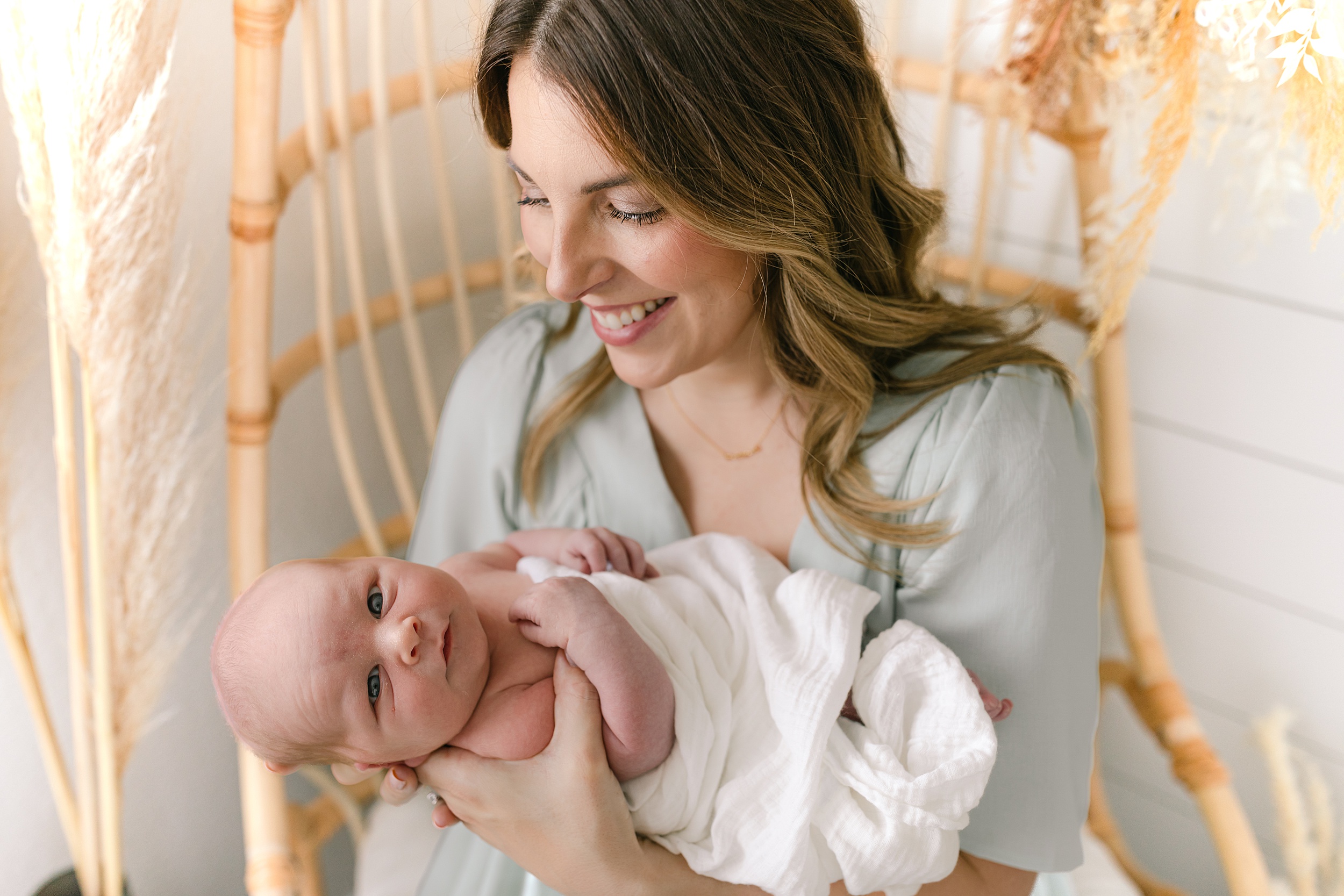 A happy mother sits in a wicker chair smiling down at her awake newborn baby in a white blanket after meeting orange county nannies