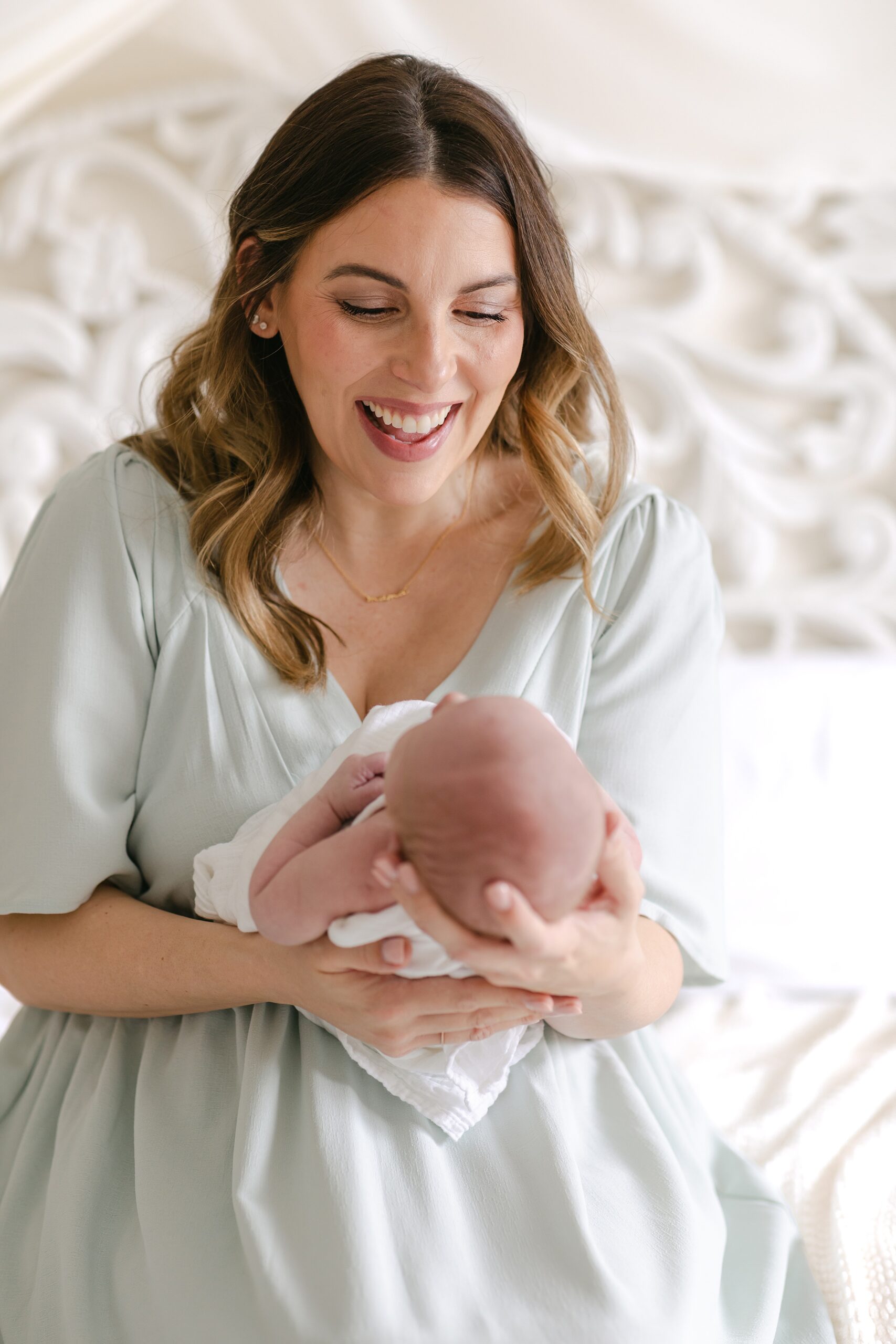 A happy mother smiles while holding her newborn baby in her hands while sitting on a bed after meeting orange county nannies