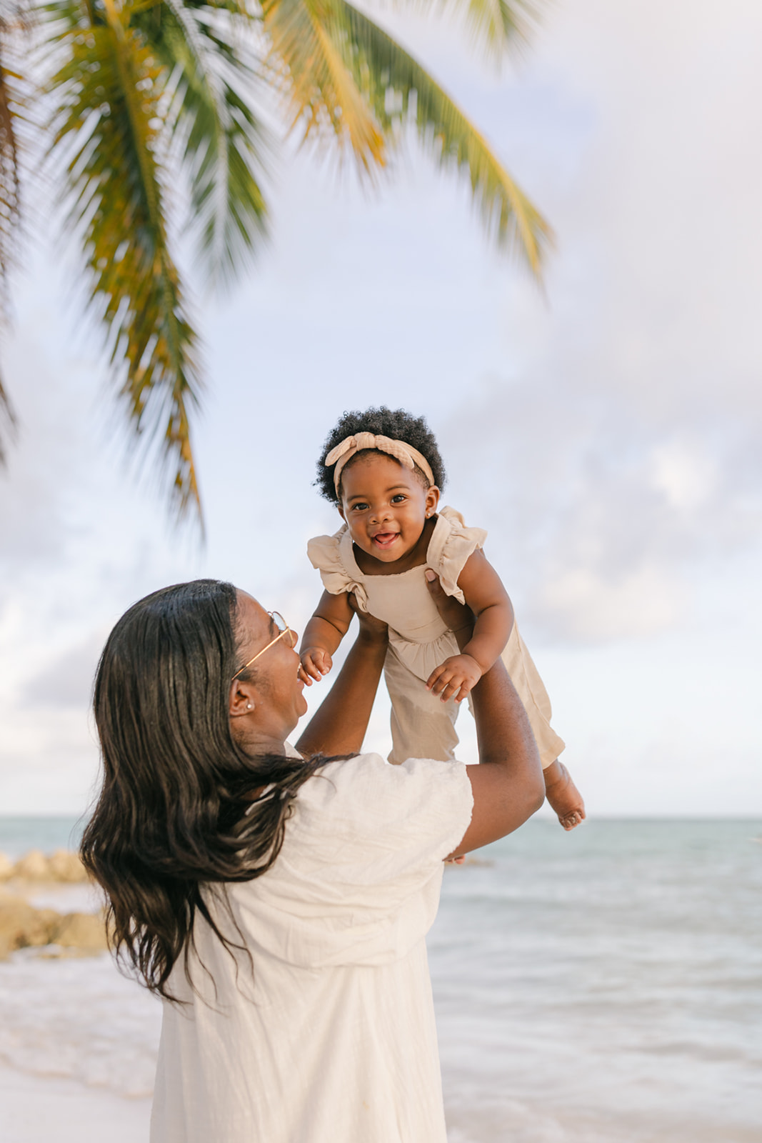 A mother in a white dress lifts her laughing toddler daughter while standing on a beach before visiting orange county Pediatrician