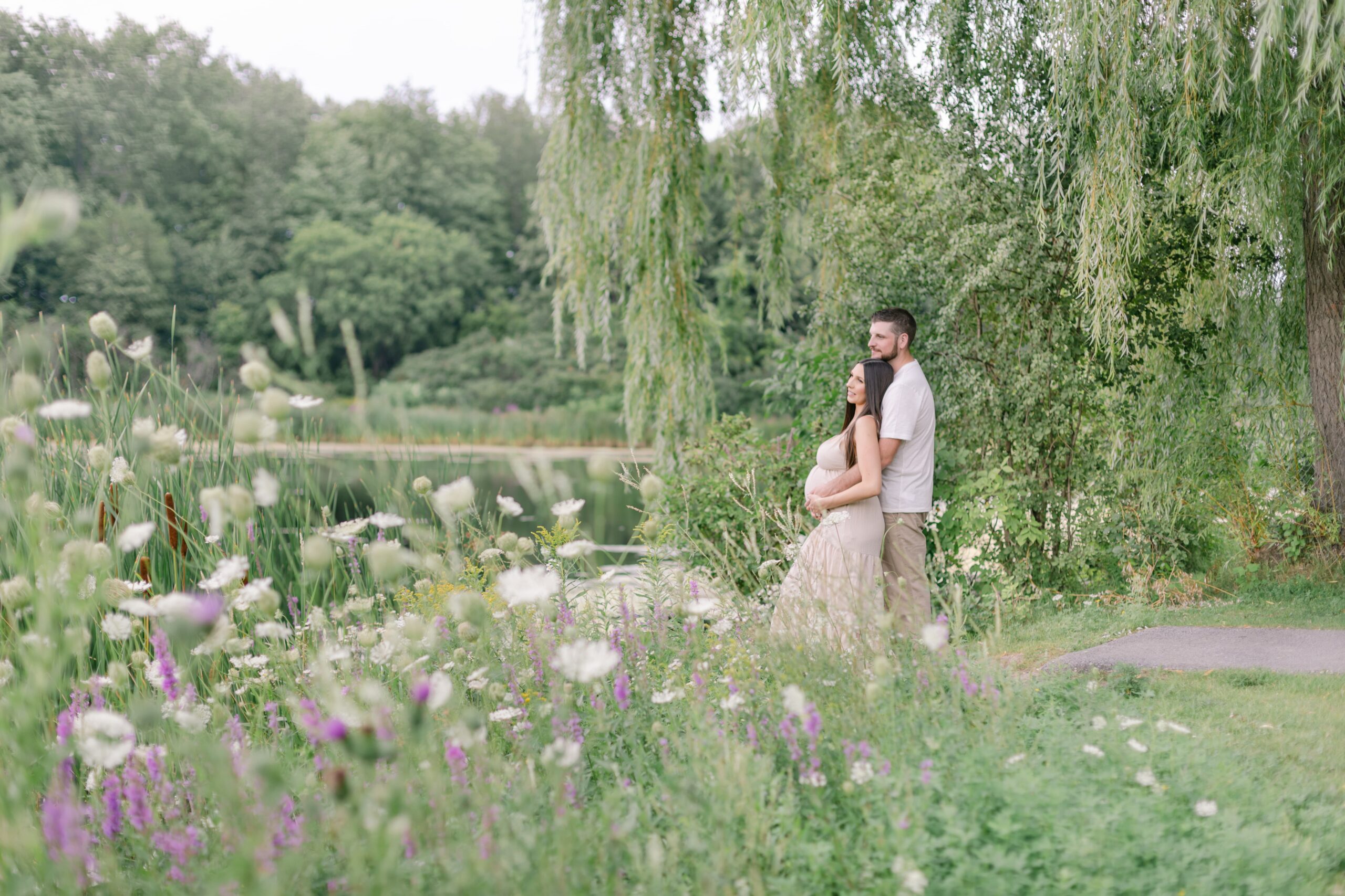 Expecting parents stand on the edge of a lake under a willow tree holding the bump