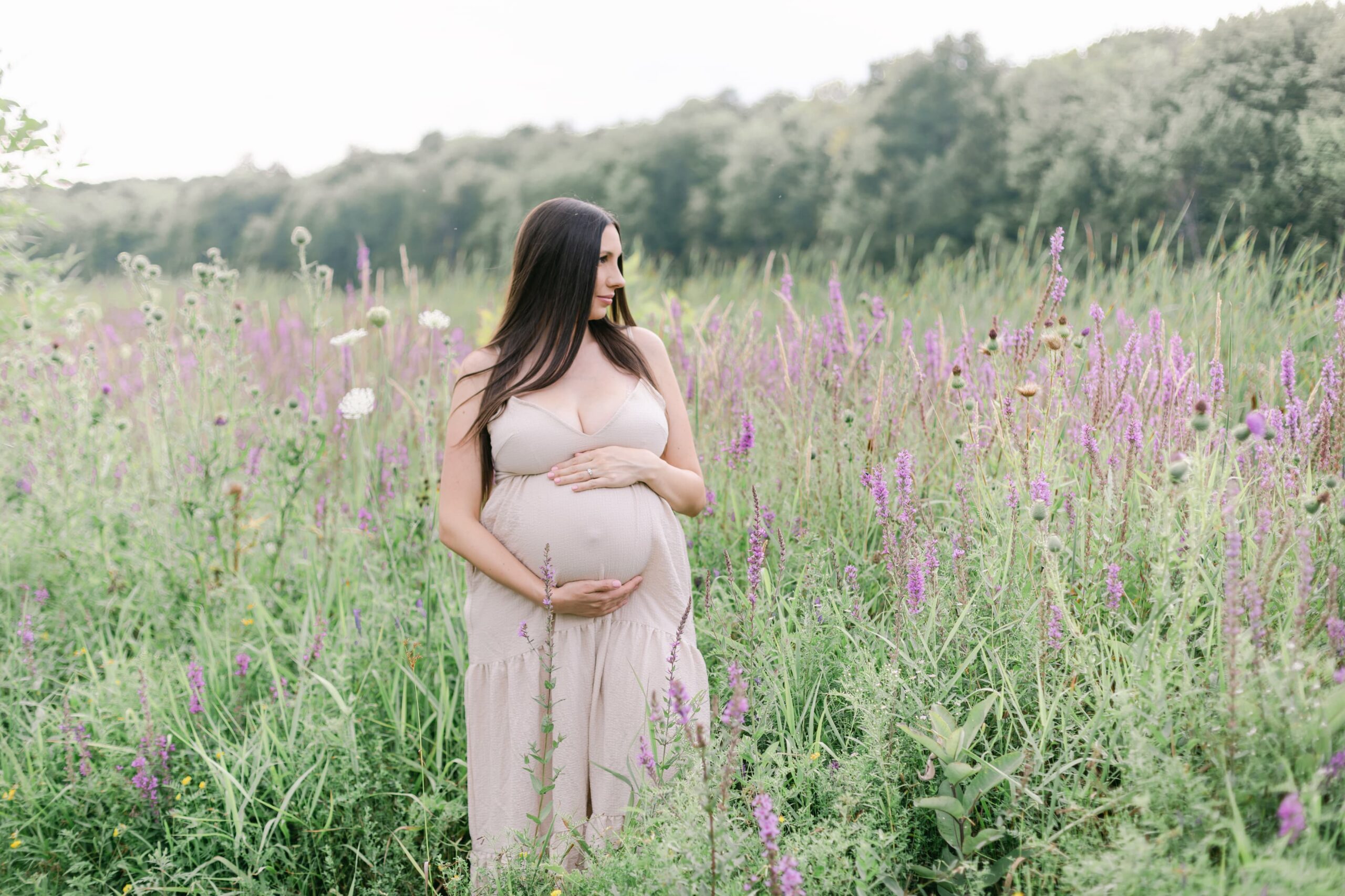 A mother to be in a beige dress holds her bump while standing in a field of wildflowers before some orange county prenatal yoga