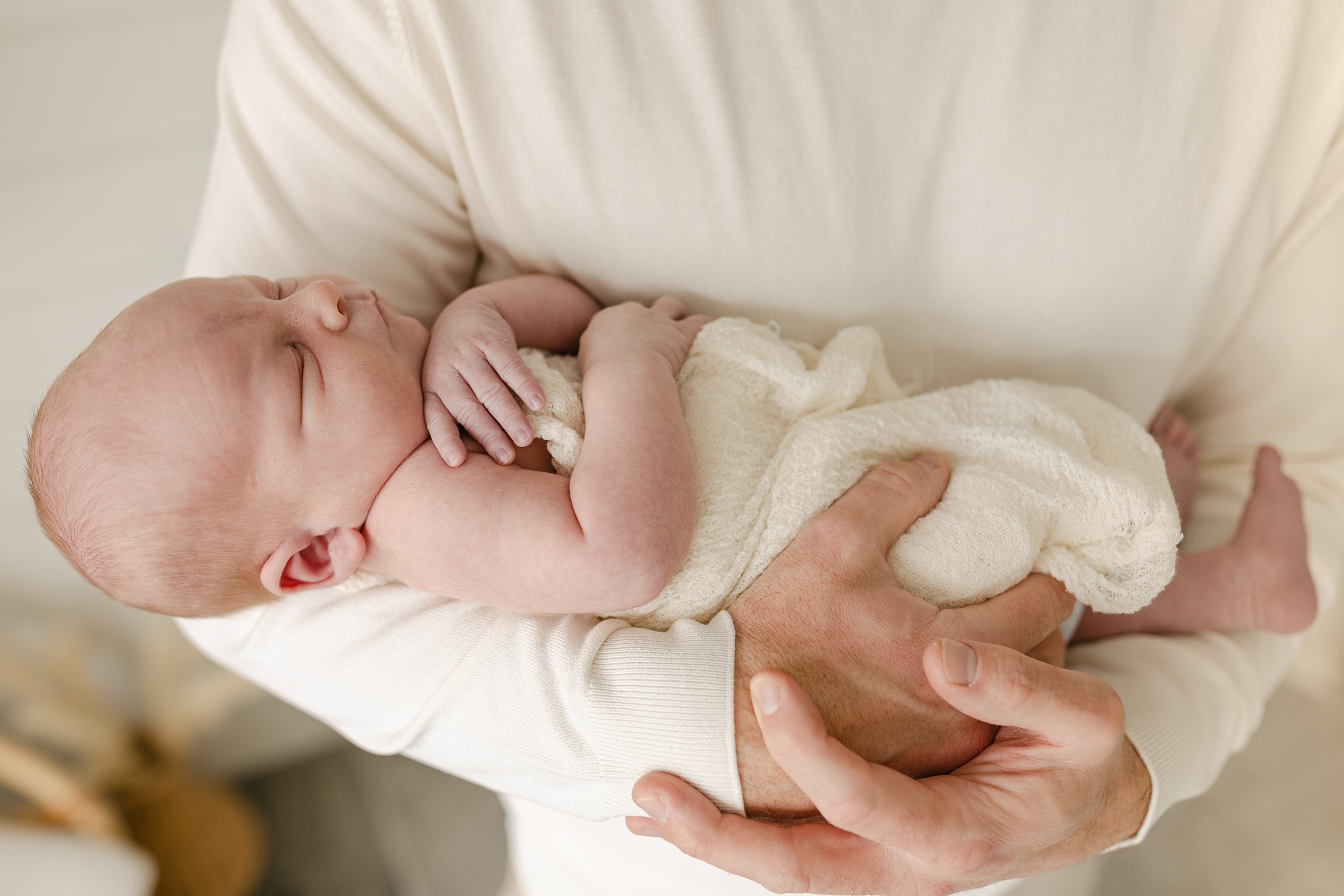 A father cradles his sleeping newborn baby wrapped in a white swaddle