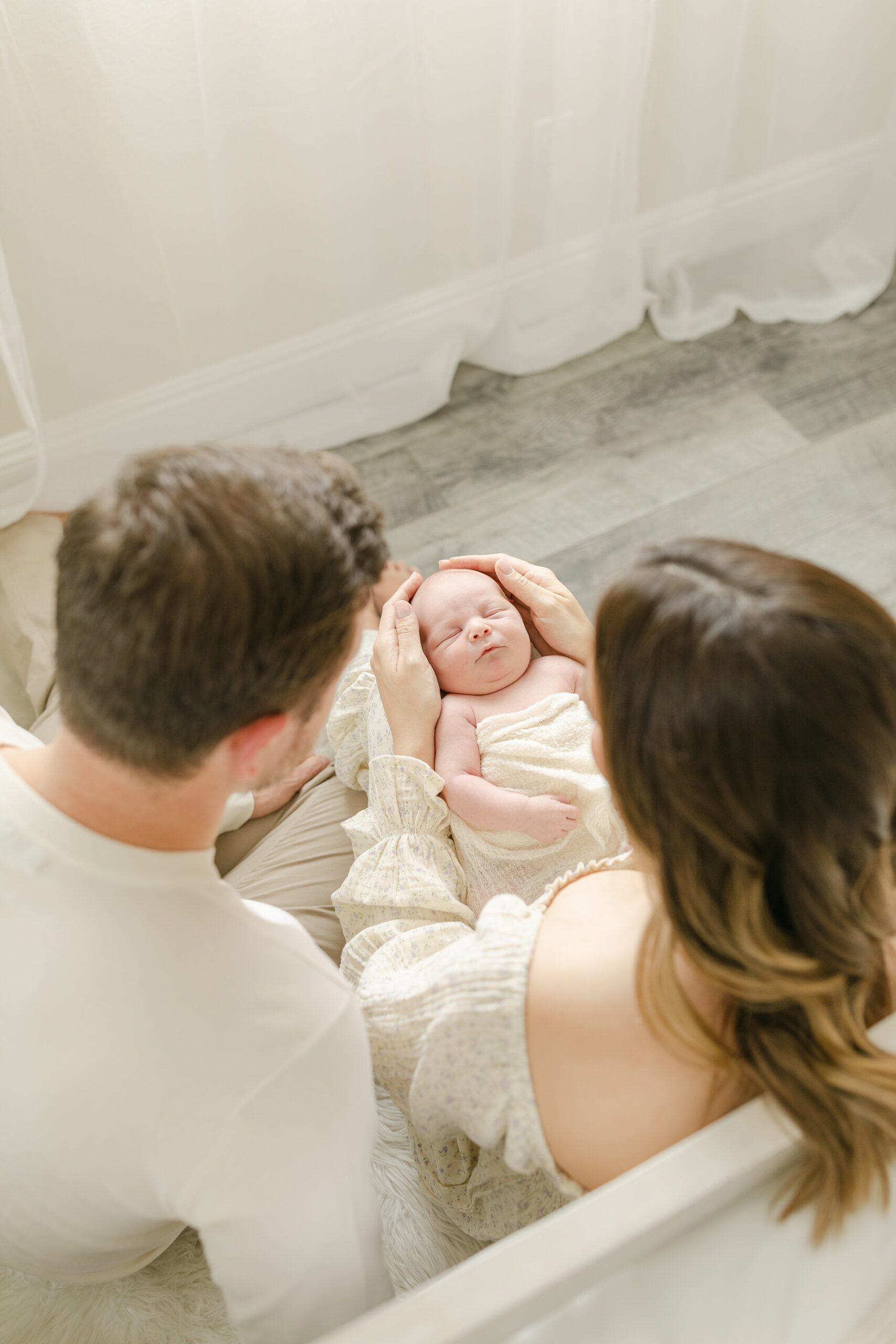 New parents look down at their newborn baby sleeping in moms lap as they sit on a couch in a studio after finding placenta encapsulation orange county