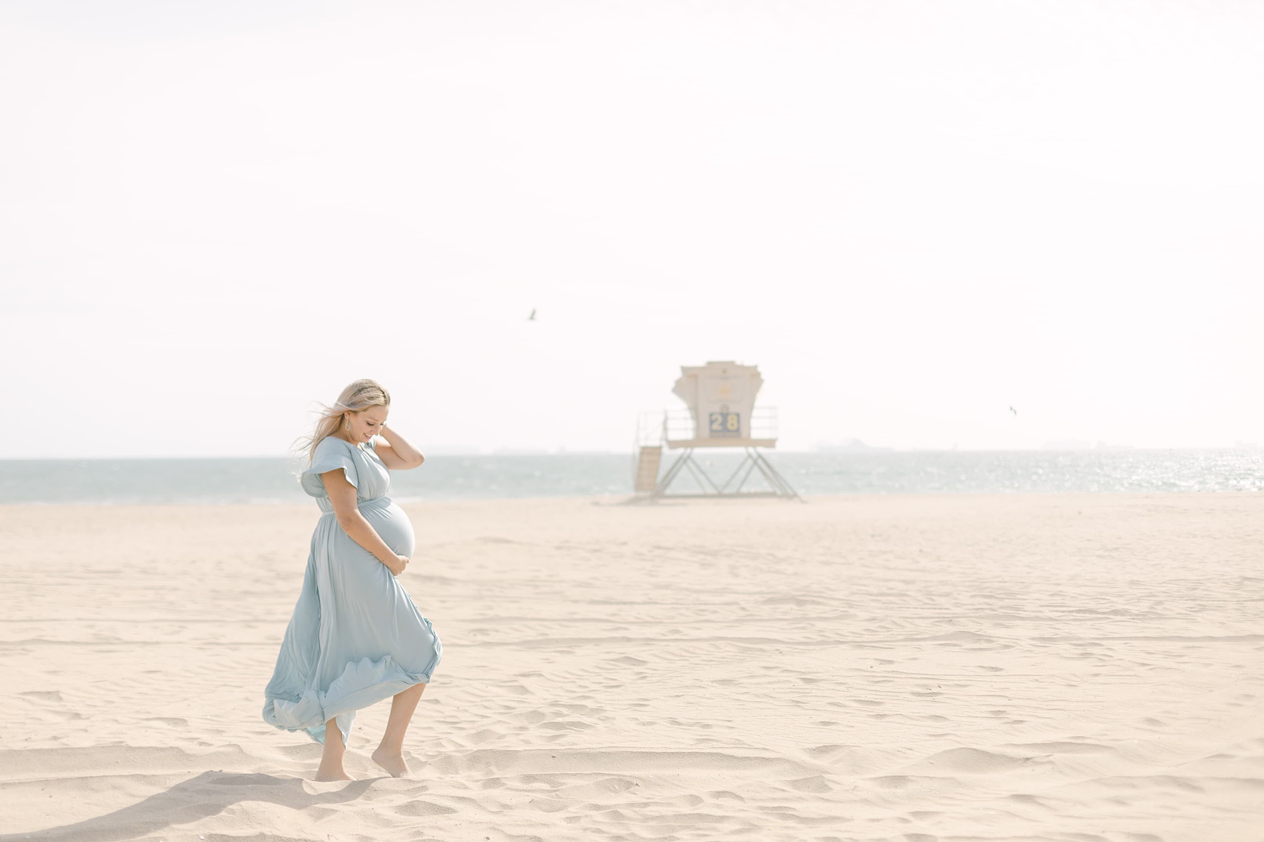 A mother to be stands on a beach in a blue maternity gown smiling down at her bump