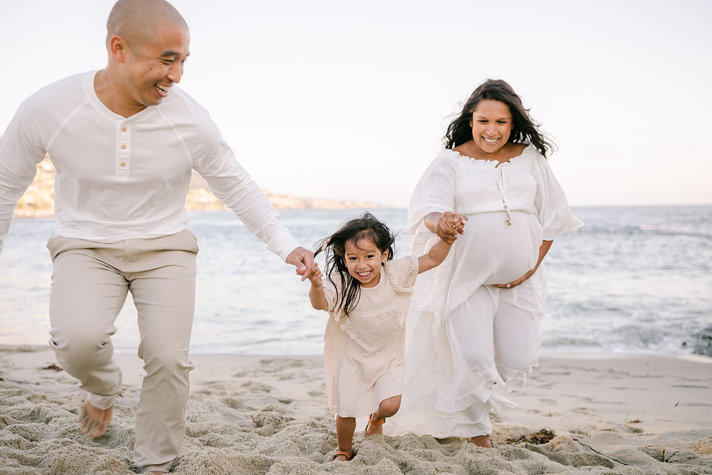 A pregnant mother in a white maternity gown walks up a beach playing with her husband and their toddler daughter after a prenatal massage newport beach