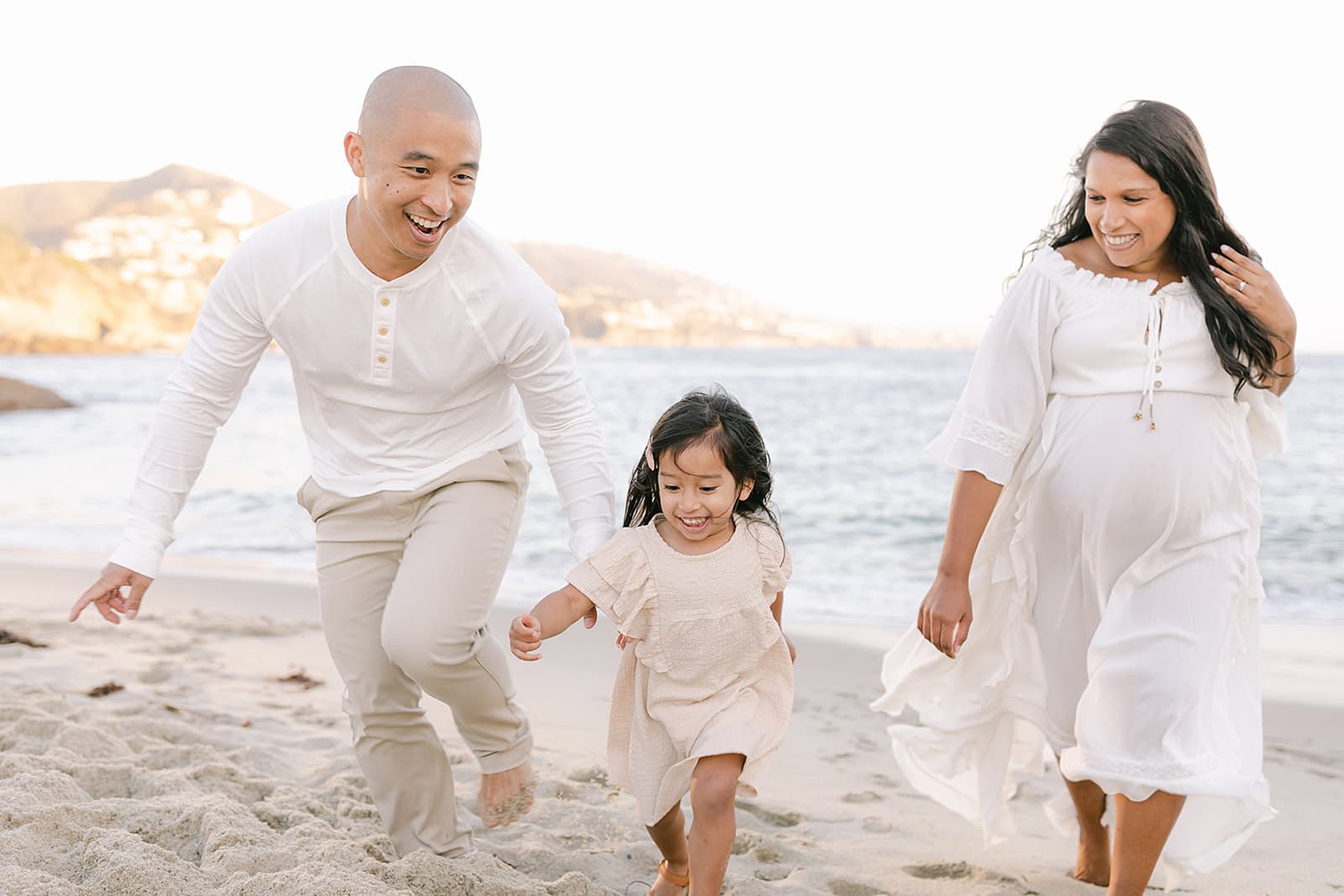 A mom and dad in white chase and play with their toddler daughter on a beach at sunset