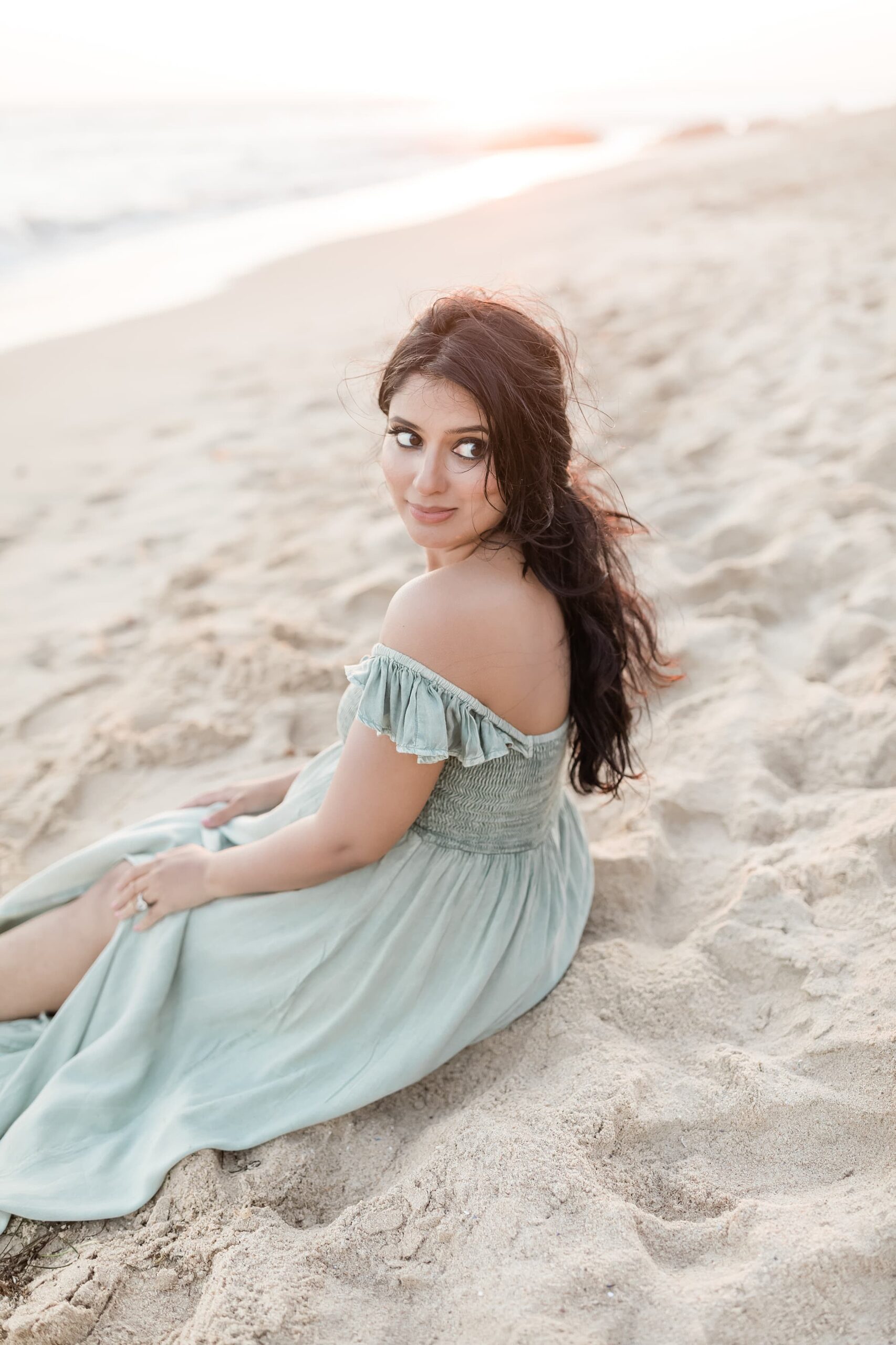 A mom to be in a green maternity dress sits on the beach while looking over her shoulder at sunset after a prenatal massage newport beach