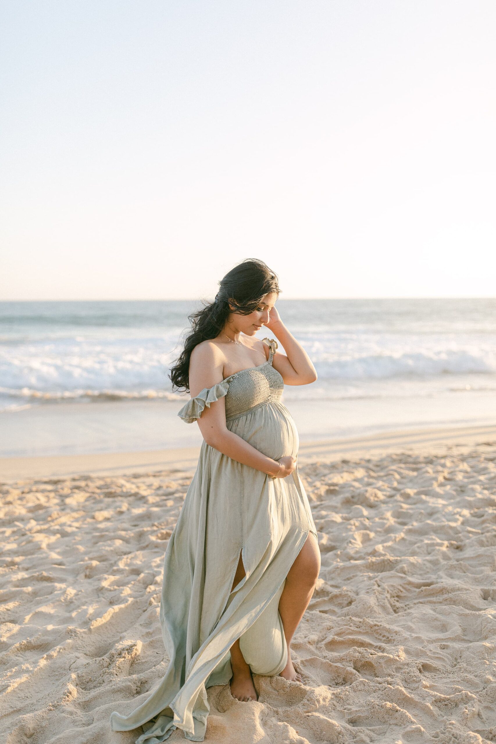 A mother to be in a green maternity dress stands on a windy beach holding her hair back and her bump after a prenatal massage newport beach