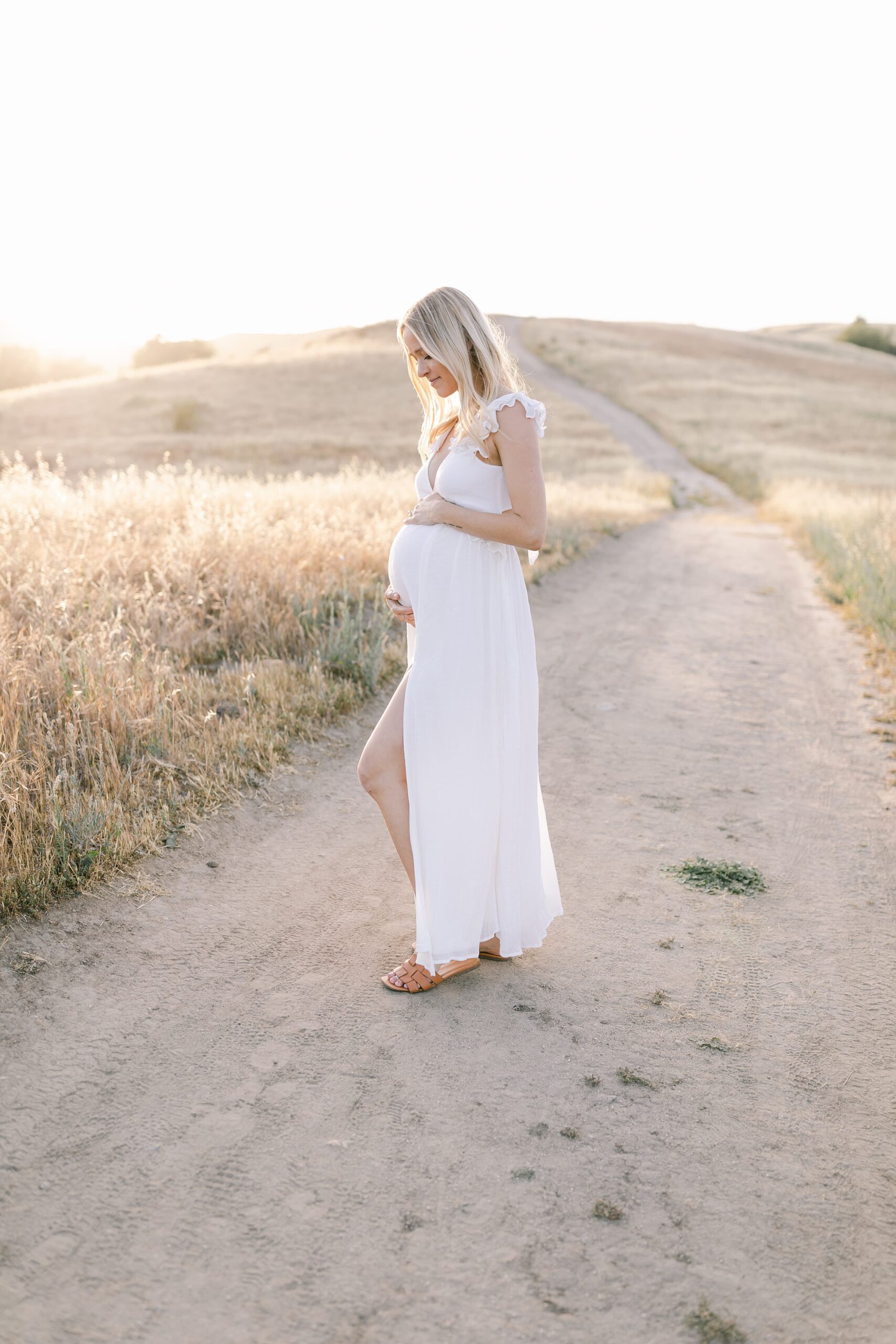 A mom to be in a white maternity dress smiles down at her bump while standing in a park trail