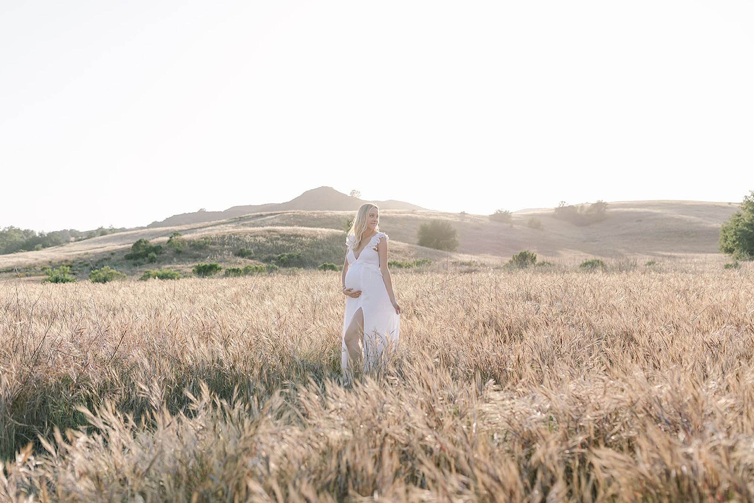 An expecting mother walks through a vast field of golden grass at sunset in a white maternity gown