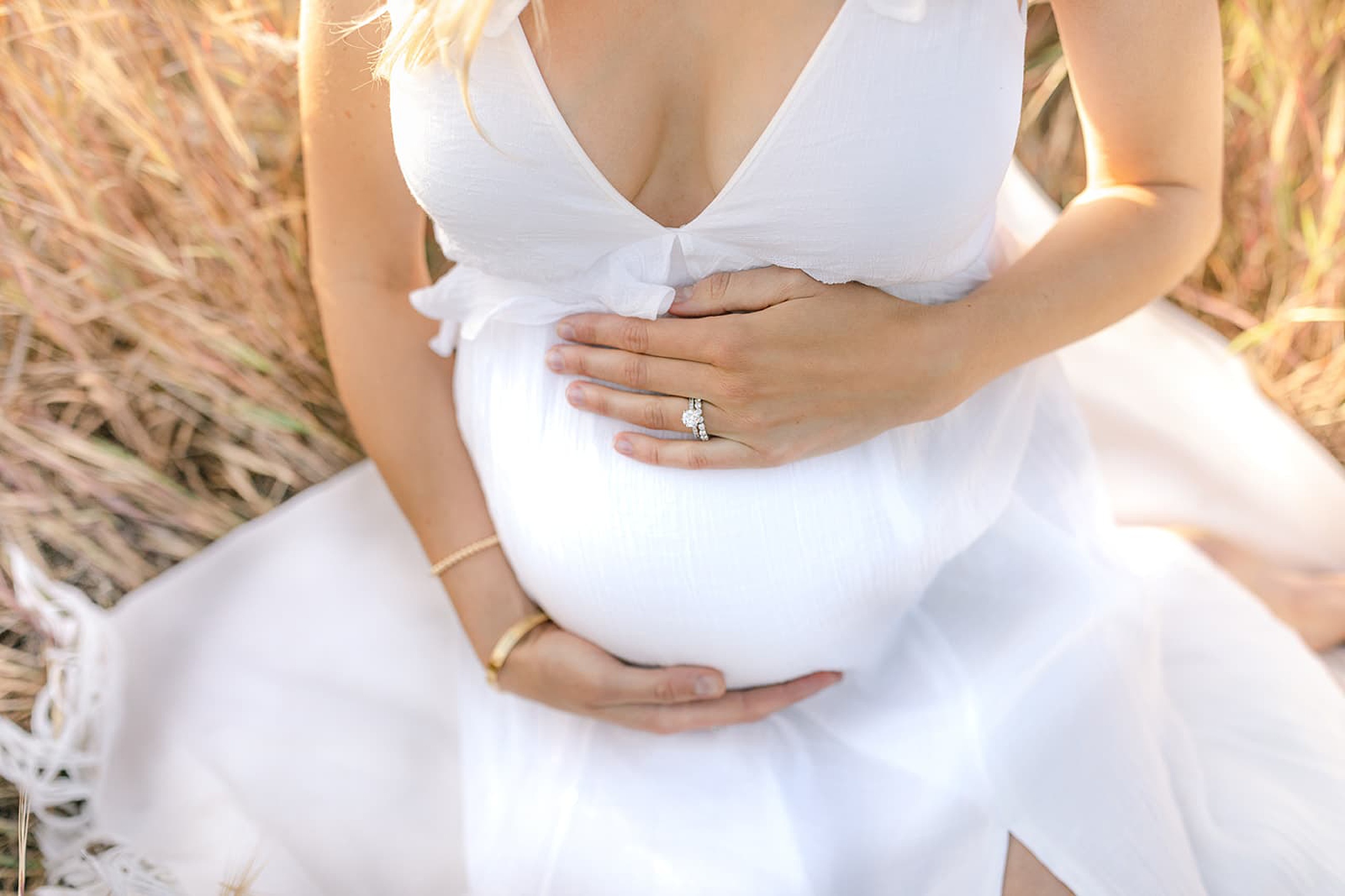 Details of a mom to be sitting in some golden grass at sunset holding her bump in a white dress after some prenatal massage orange county