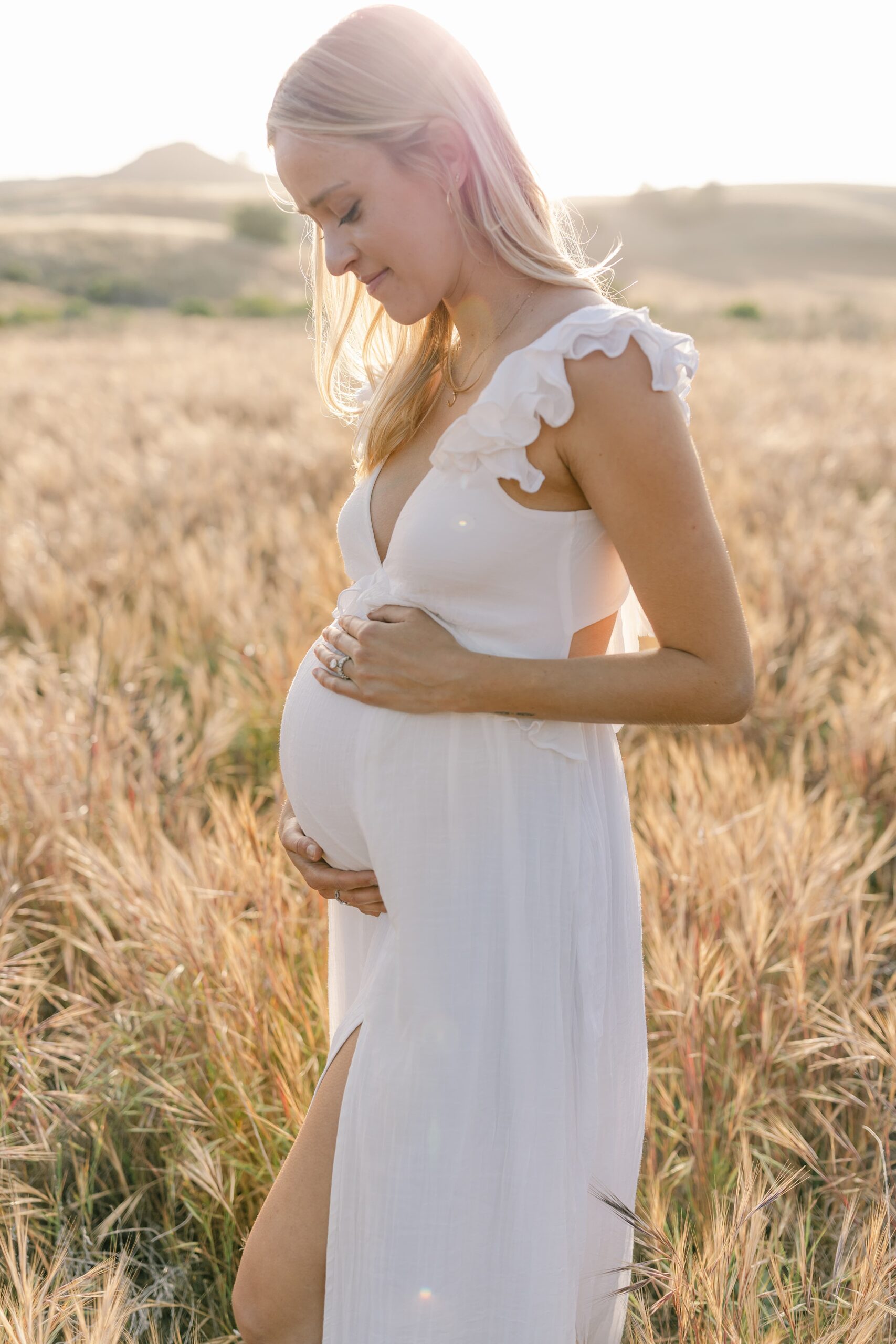 A mother to be in a white maternity gown stands in a field of tall golden grass smiling down at her bump at sunset after a prenatal massage orange county