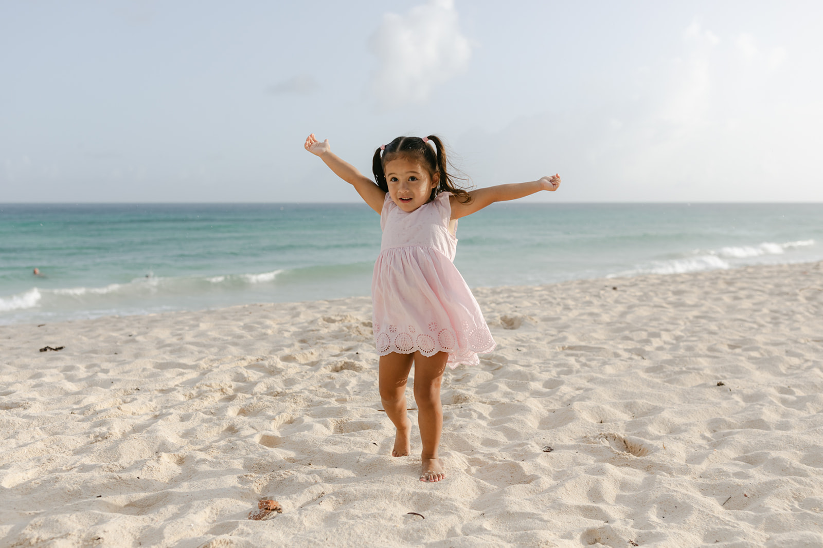 A toddler girl in a pink dress plays in the sand on a beach