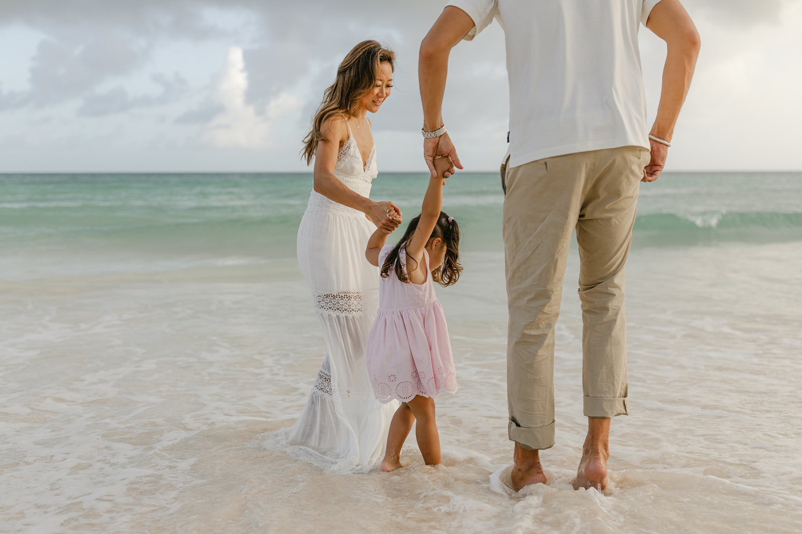 A mom and dad walk their toddler daughter by the hand through the water on a beach