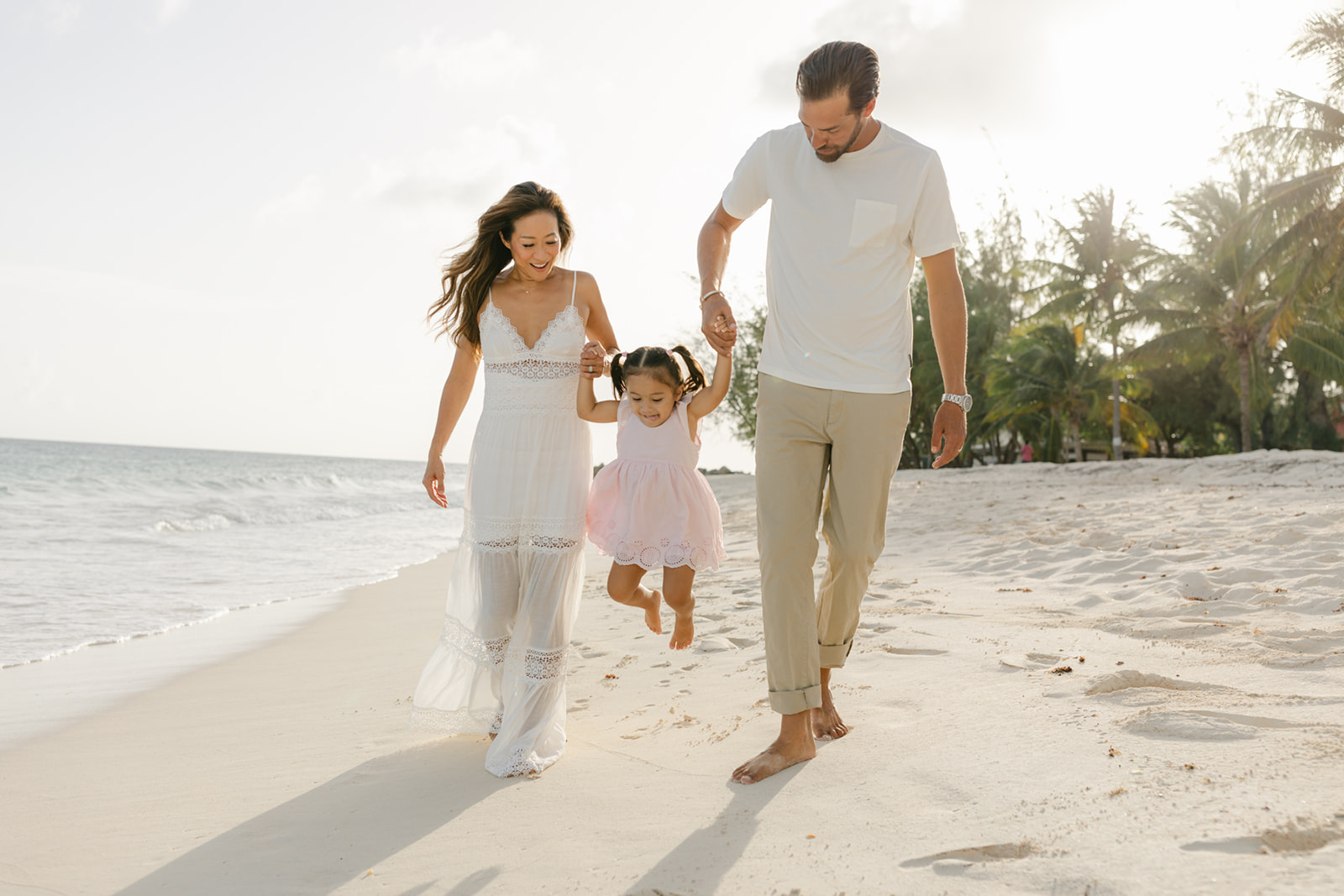 Happy parents walk their toddler daughter down a beach in a pink dress and lift her during things to do in newport beach with kids