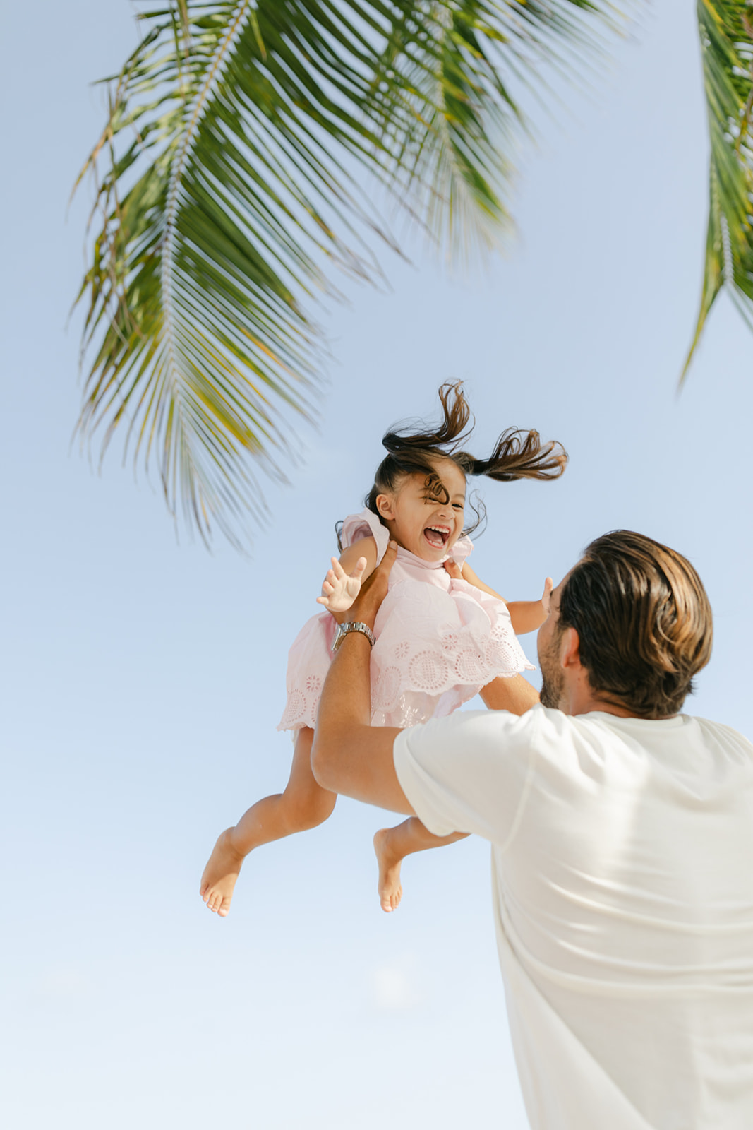 A dad in a white shirt lifts his toddler daughter up in the air as she laughs in a pink dress on a beach during things to do in newport beach with kids