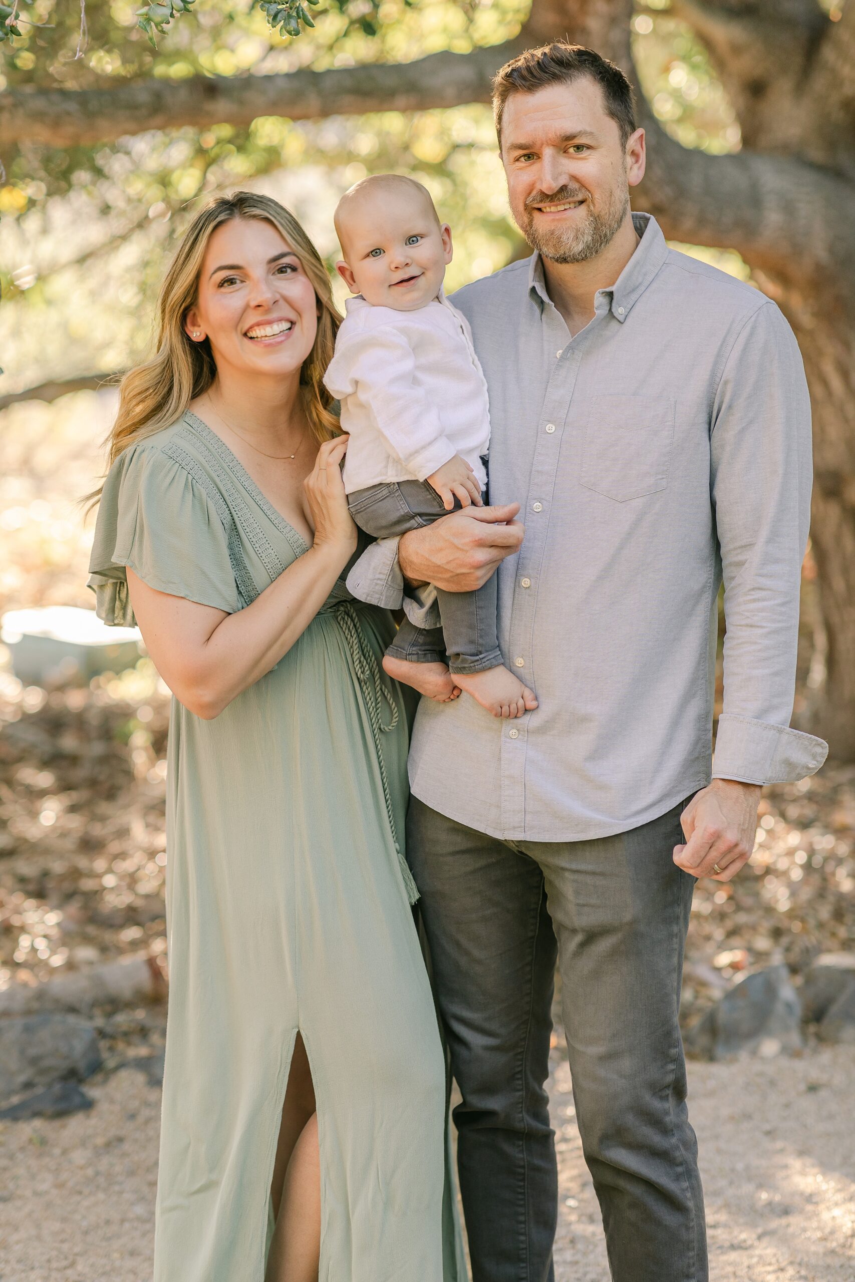 Happy parents stand smiling with their infant son in dad's arm in a park at sunset