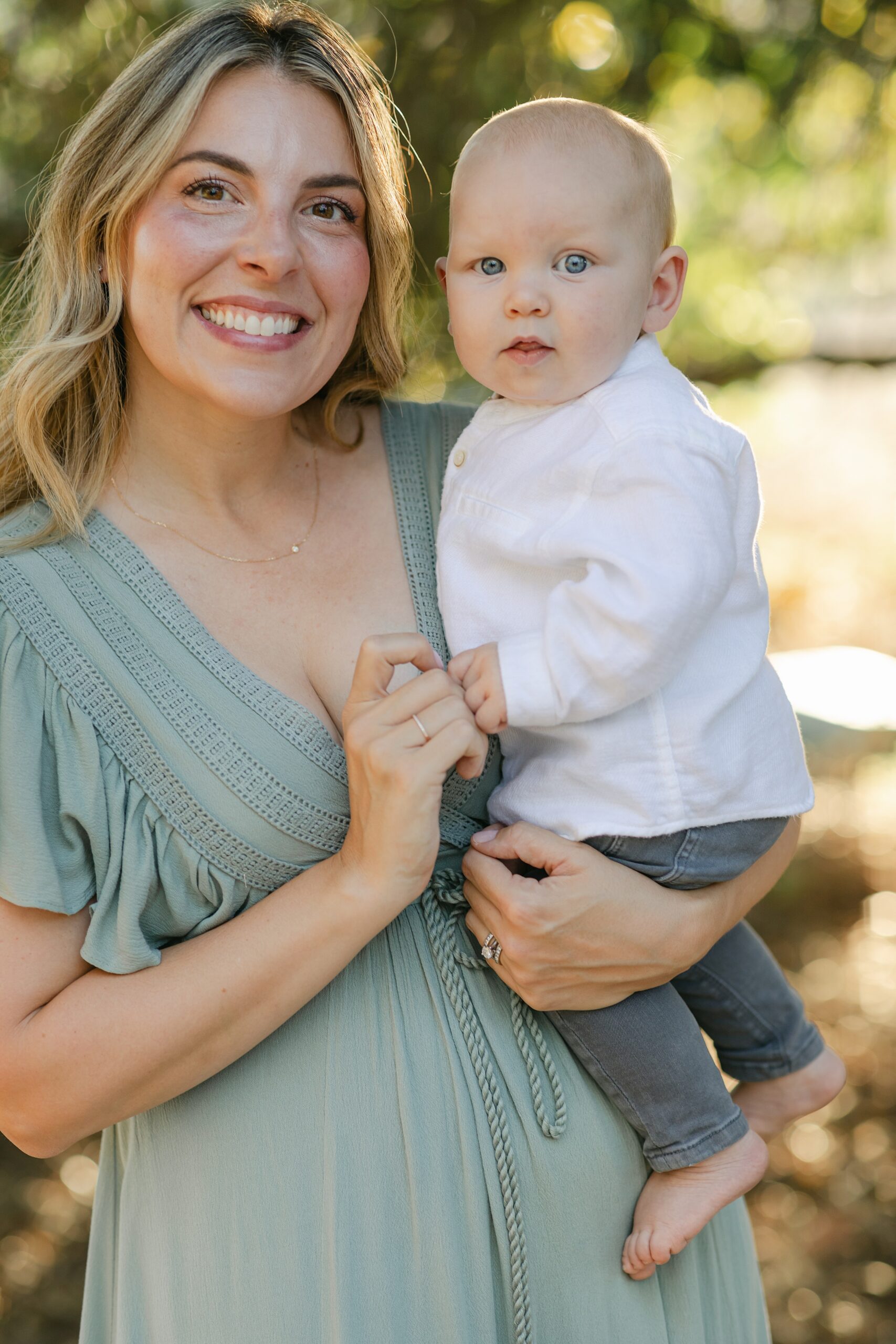 A happy mom in a green dress stands under some trees smiling with her infant son