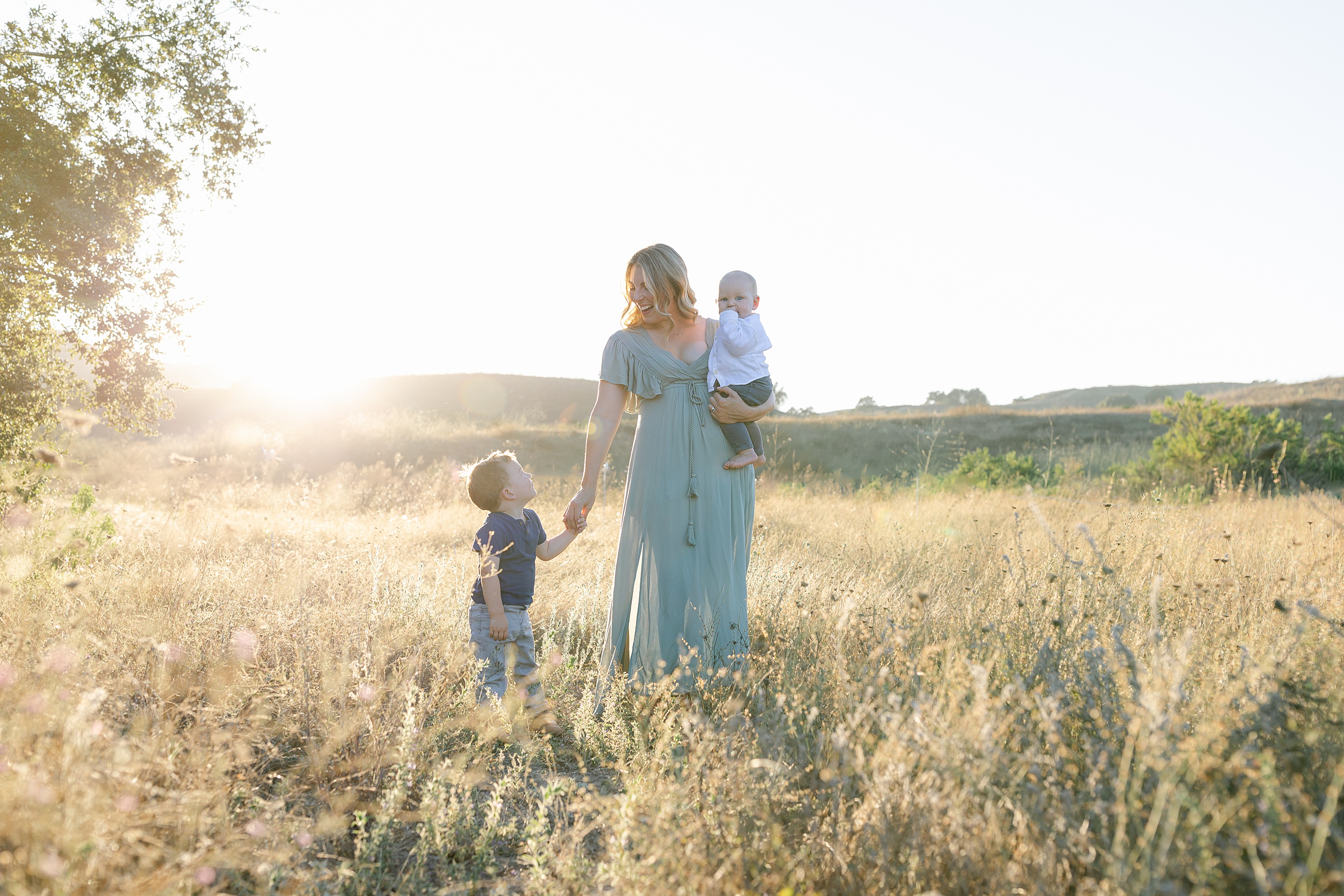 A happy mom smiles while holding her infant son on her hip and holding the hand of her toddler son at her side while walking through a field of tall golden grass at sunset during things to do in orange county with toddlers