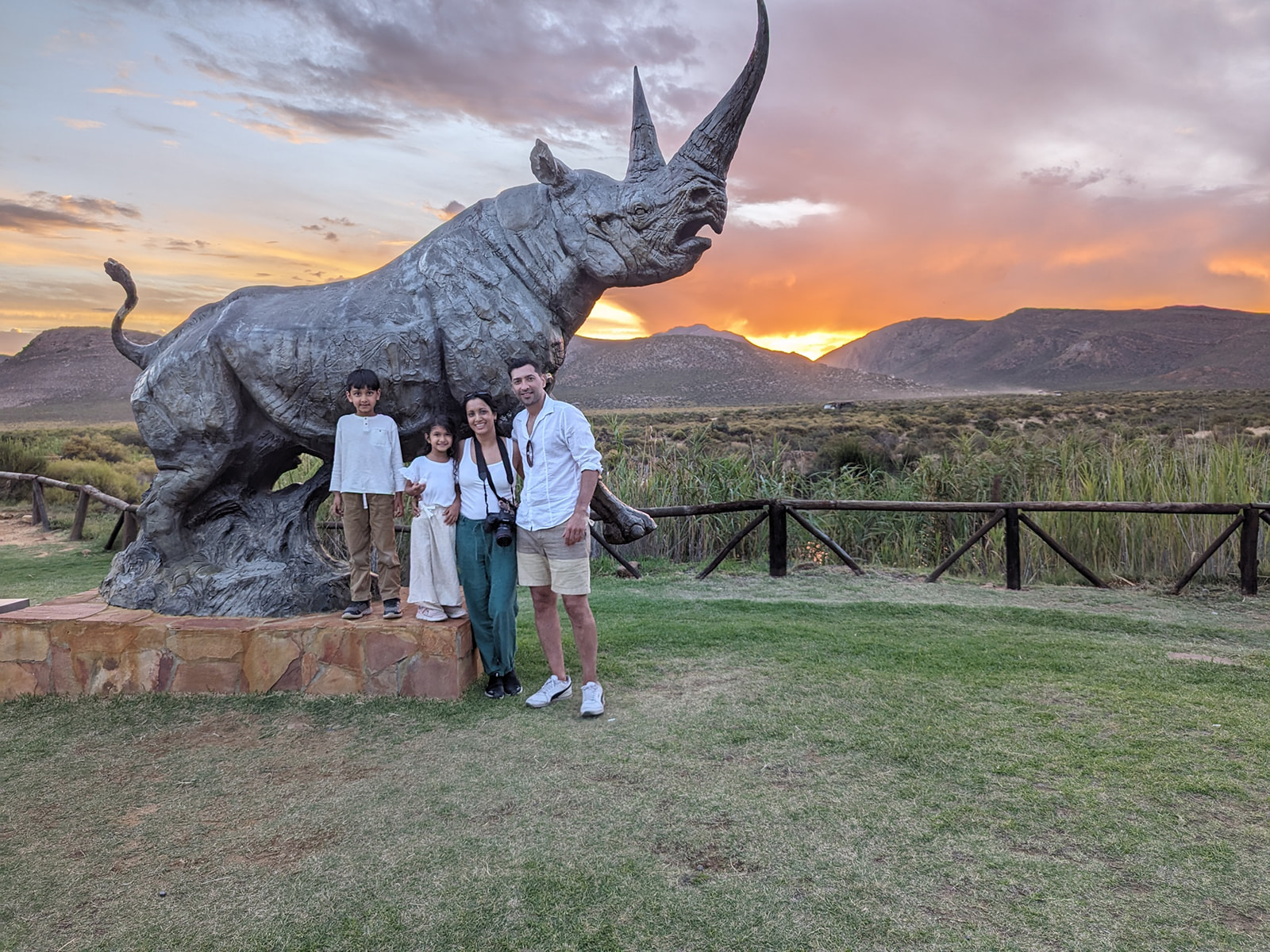 The Sondangar family poses by a hippopotamus statue in South Africa with a stunning sunset taking place in the mountains behind. s